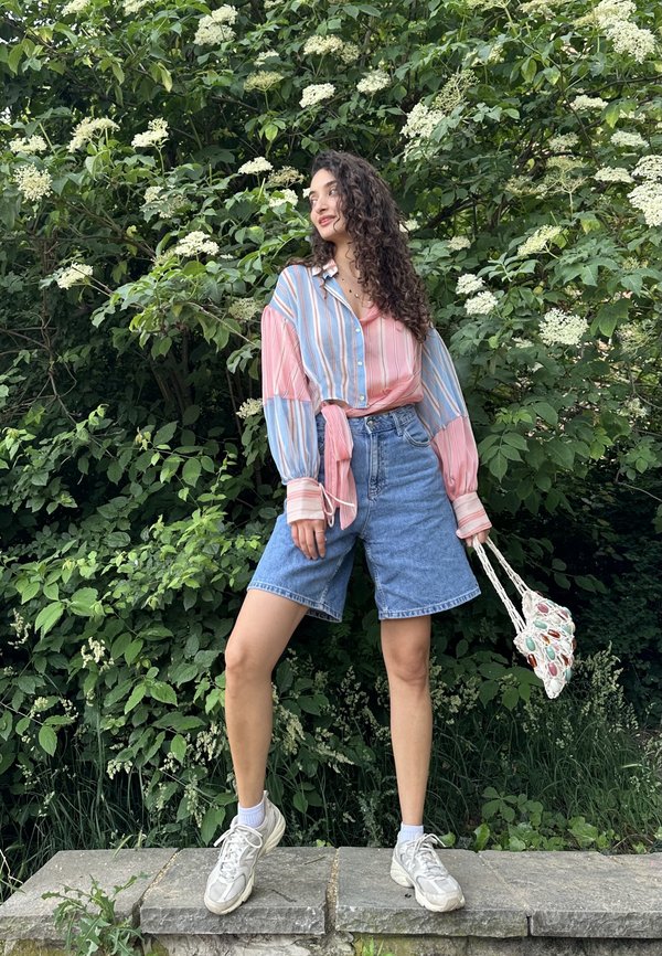 Kvinde med langt krøllet hår står på en stenforkant foran grønne buske med hvide blomster iført en stribet skjorte og denimshorts.
