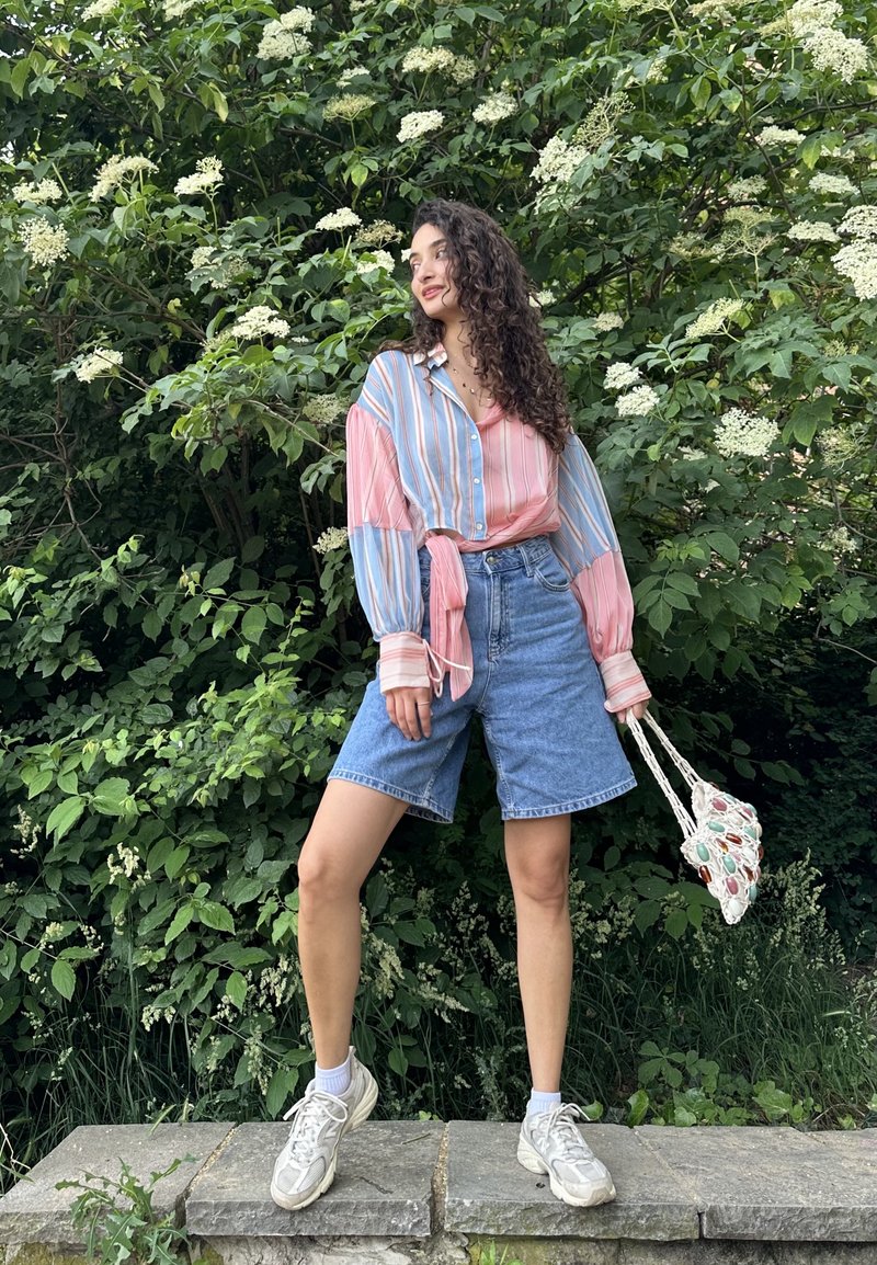 Femme aux longs cheveux bouclés se tenant sur une corniche en pierre devant des buissons feuillus verts avec des fleurs blanches, portant une chemise rayée et un short en jean.