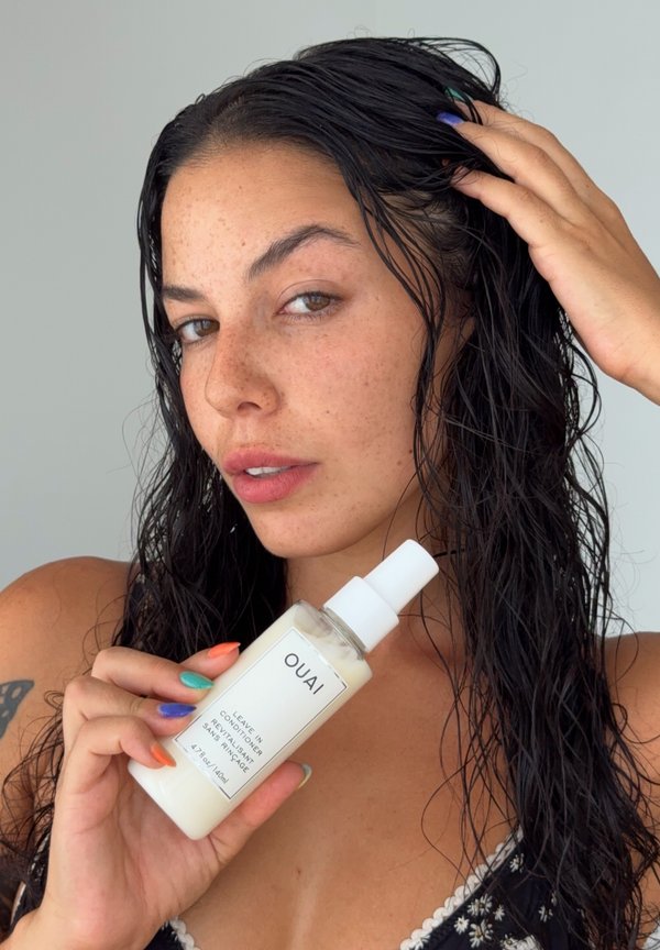 Vrouw met nat donker haar houdt een fles OUAI leave-in conditioner dicht bij haar gezicht terwijl ze met haar andere hand haar haar aanraakt.