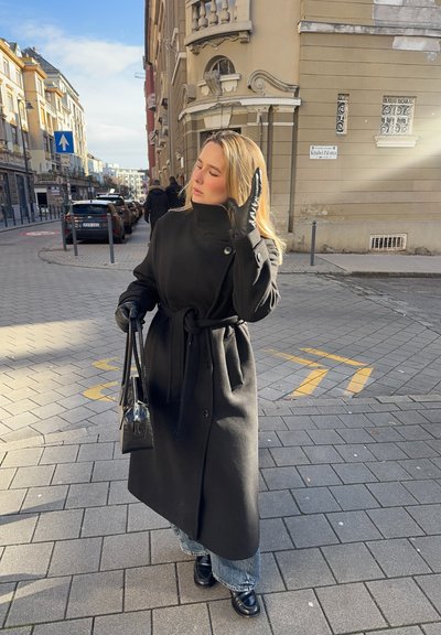 Abrigo negro con cinturón hecho de lana, con cuello alto y detalles de botones. Combina con jeans azul claro y botas negras. Cielos despejados arriba.