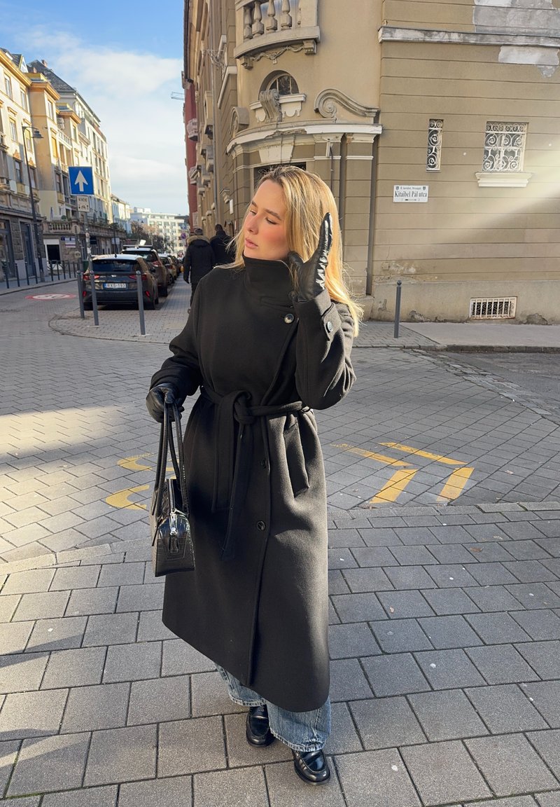 Schwarzer Wollmantel mit Gürtel, hohem Kragen und Knopfdetails. Kombiniert mit hellblauen Jeans und schwarzen Stiefeln. Klarer Himmel darüber.