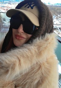 Personne portant de grandes lunettes de soleil noires, une casquette beige, une capuche sombre et un épais manteau en fourrure beige, debout à l'extérieur avec un paysage urbain enneigé en arrière-plan.