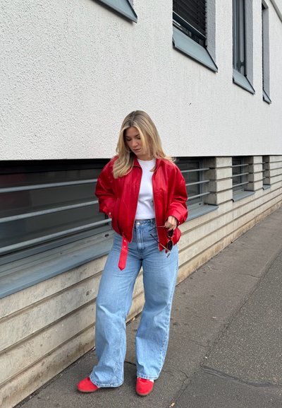 Joven mujer con chaqueta y zapatos rojos, con camisa blanca y vaqueros azules, de pie en la acera cerca de un edificio moderno con ventanas horizontales.