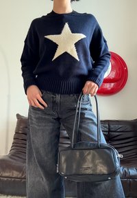 Pull bleu marine avec un motif étoile blanche, poignets côtelés, associé à un jean gris foncé à jambes larges. Tient un sac à main en cuir noir avec une fermeture éclair.