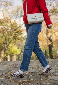 Személy kék farmert, piros pulóvert, fehér kézitáskát, fehér zoknit és leopárdmintás cipőt viselve sétál macskaköves úton őszi környezetben.