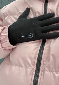 Luvas de malha pretas com o logótipo branco da Nike, repousando contra uma jaqueta puffer rosa com fecho éclair à frente e detalhes de punho franzido.