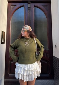 Femme portant un pull vert, une jupe blanche en dentelle superposée, un foulard fleuri, des lunettes teintées vertes et plusieurs colliers, debout devant une porte en bois.