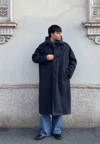 Άνδρας με μακρύ μαύρο παλτό και κουκούλα στέκεται μπροστά σε γκρι τοίχο, κοιτώντας πλάγια με το ένα χέρι μέσα στην τσέπη του παλτού και το άλλο κοντά στο στήθος.