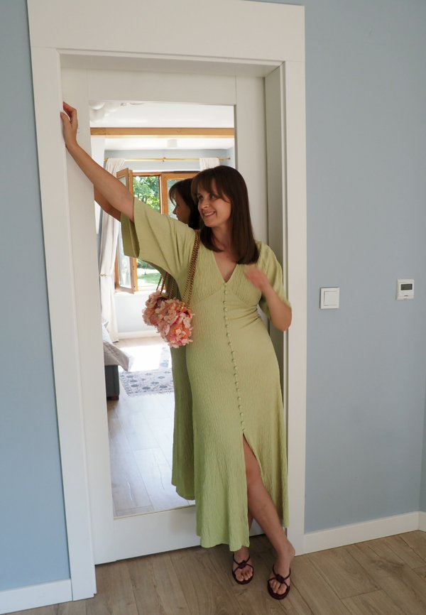 Femme en robe vert clair appuyée contre un cadre de porte, tenant un sac à fleurs, debout pieds nus sur un sol en bois dans une pièce lumineuse.