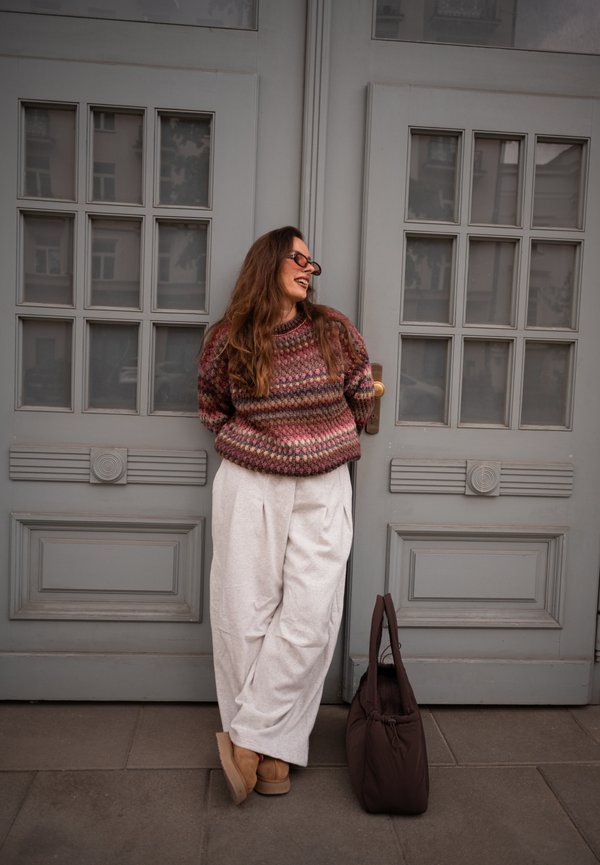 Femme aux longs cheveux portant un pull à motifs, un pantalon blanc et des lunettes de soleil, appuyée contre une porte double grise avec un sac sombre posé au sol à proximité.