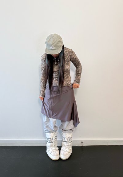 Top de manga larga con estampado de camuflaje combinado con una falda marrón, pantalones jogger grises y zapatillas altas blancas. Una gorra gris claro completa el conjunto.