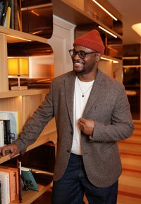 Personne portant un blazer marron à carreaux sur une chemise blanche et un jean bleu, souriant à côté d'une bibliothèque en bois avec des livres et une lampe.