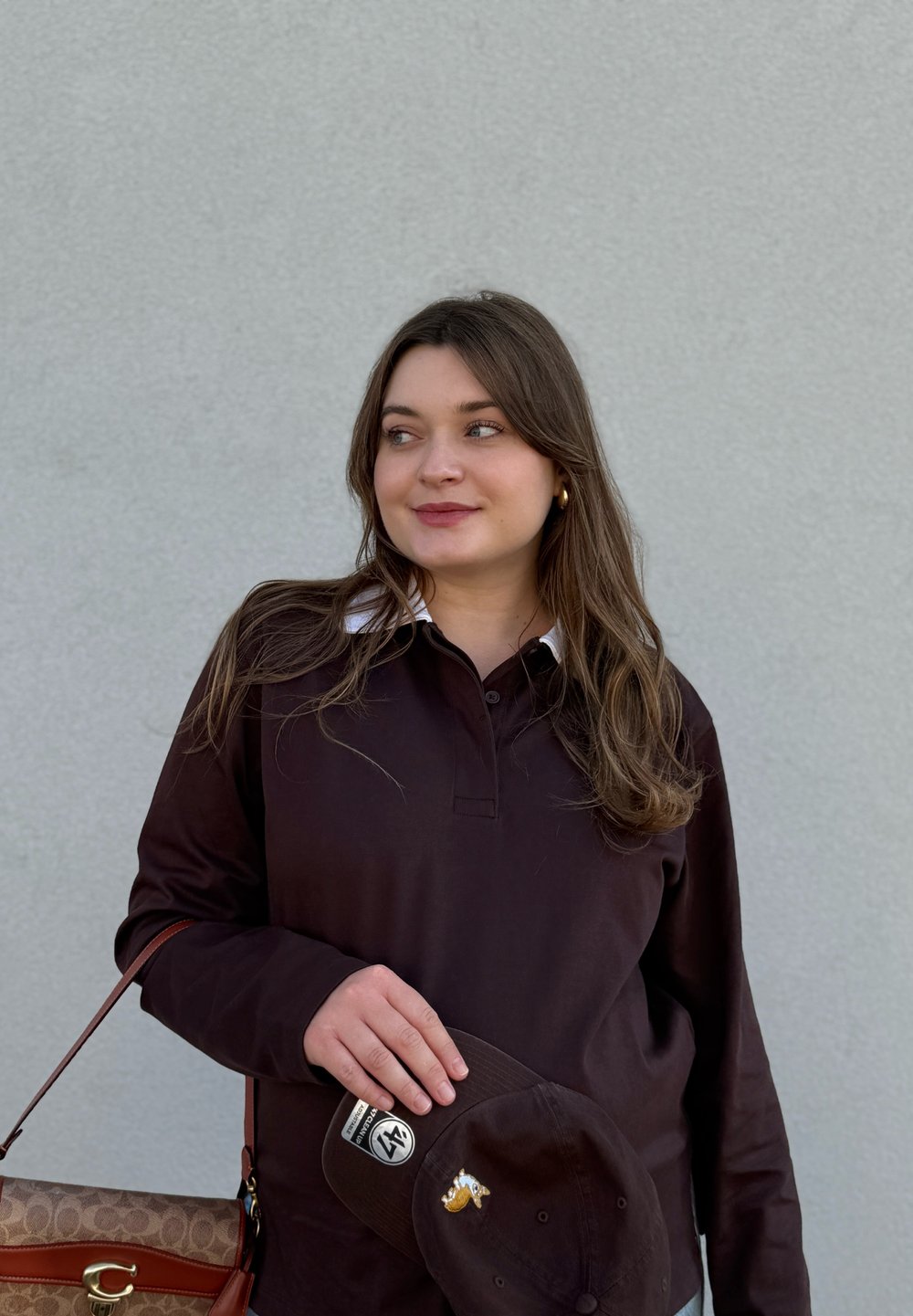 Jonge vrouw in een donker shirt met lange mouwen, die een donkere pet met patches en een bruine handtas vasthoudt, staand tegen een effen grijze muur.