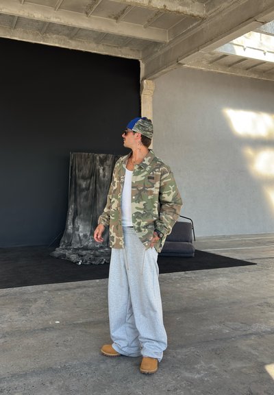 Chaqueta de camuflaje, camiseta blanca sin mangas, pantalones de chándal grises, zapatos marrones. El fondo presenta una pared oscura y tela caída en el suelo.