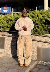 Bēšas, vaļīgas bikses, svītrains garroku krekls un saulesbrilles. Aksesuāri ietver atslēgu piekariņus, kas piestiprināti pie biksēm. Fonā redzama zaļā apstādījumi.