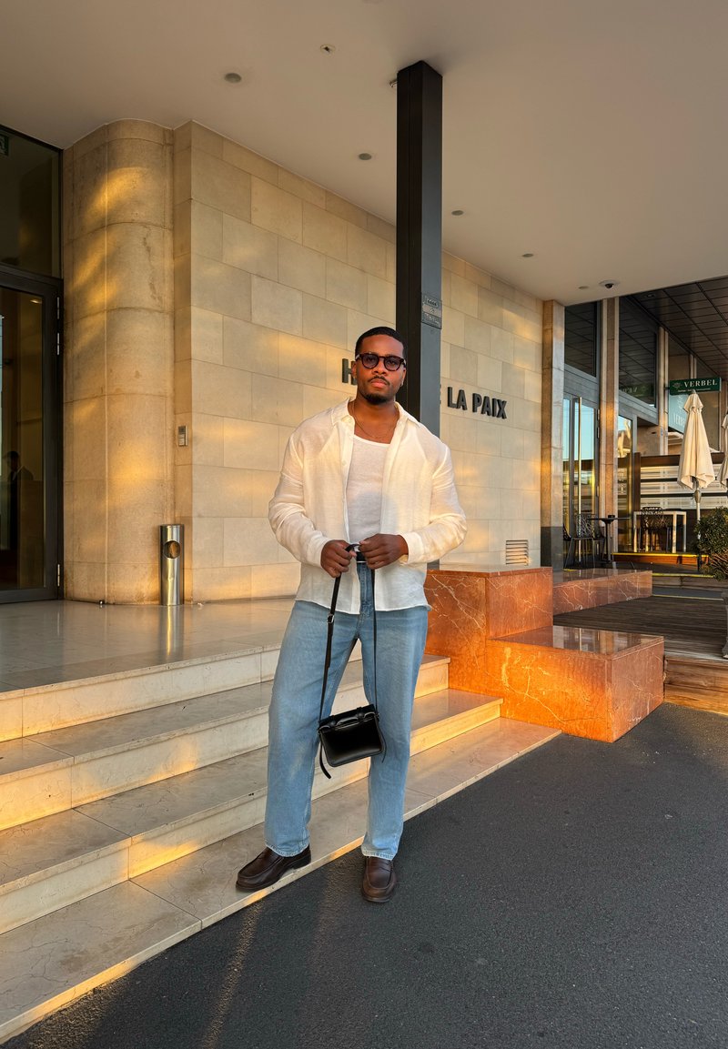 White linen shirt, white tank top, light blue jeans, and brown shoes. Holds a small black crossbody bag. Stone steps and building in background.