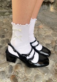 Μαύρα παπούτσια από πατίνα με τακούνια, τετραγωνισμένες μύτες και δύο λουριά στον αστράγαλο. Φορεμένα με λευκές ριγέ κάλτσες που έχουν φρουτώδεις άκρες και λεπτομέρειες από φιόγκους.