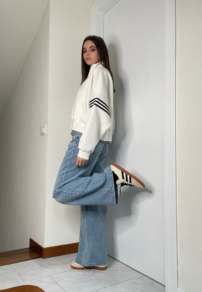 Sudadera corta blanca con rayas negras, jeans acampanados de color azul claro y zapatillas negras y blancas con suela de goma; posando contra una pared.