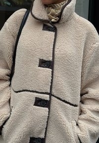 Bézs polárdzseki sötét szegéllyel, magas gallérral, elöl gombokkal és oldalzsebekkel; puha tapintású és bő szabású.