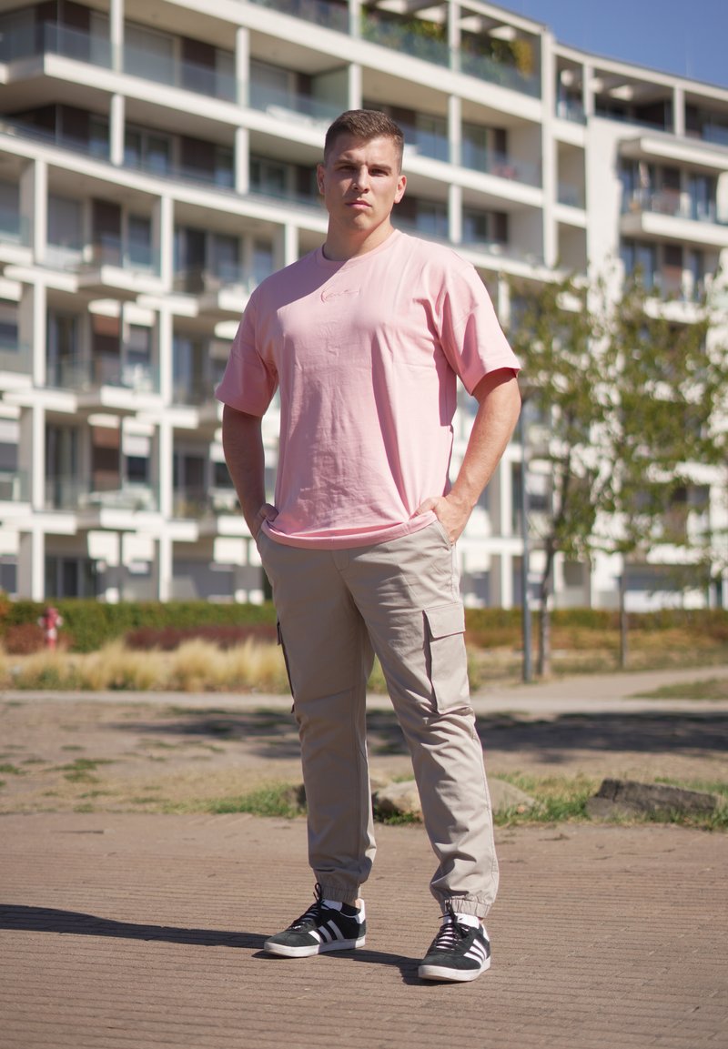Jeune homme debout à l'extérieur portant un t-shirt rose pâle, un pantalon cargo beige et des baskets noires devant un immeuble d'appartements.