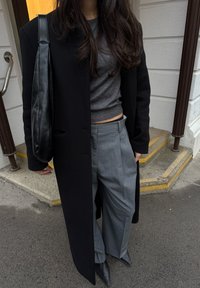 Manteau en laine noir, haut en maille gris, pantalon large gris, chaussures noires pointues et sac en cuir noir. Design simple avec des couleurs sobres et des lignes épurées.