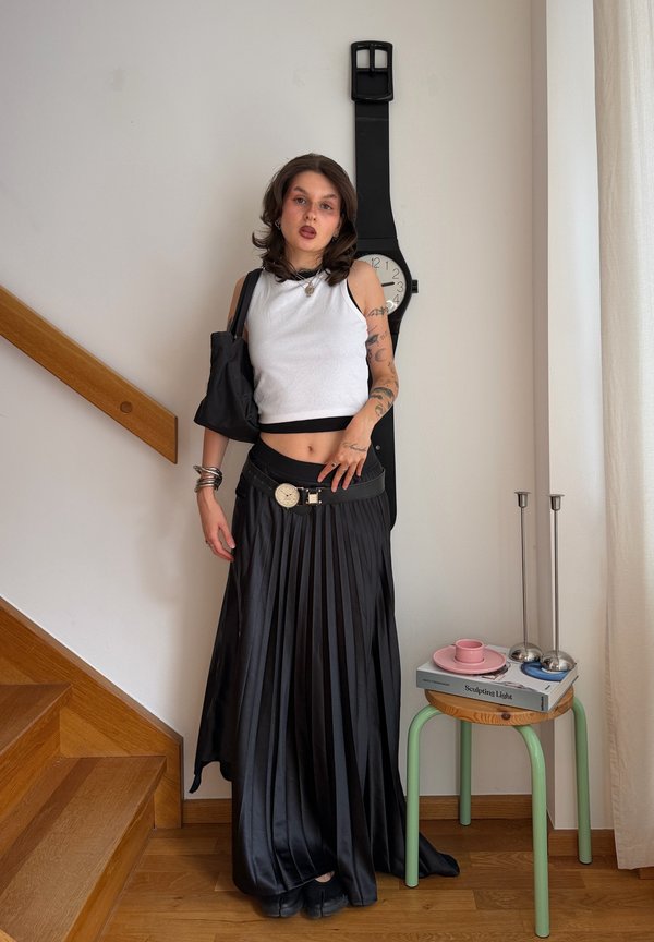 Giovane donna con tatuaggi indossa un top bianco senza maniche e una gonna nera plissettata con cintura a orologio, in piedi vicino a un grande orologio da parete e scale di legno.