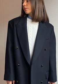 Γυναίκα με μαλλιά έως τους ώμους, φορώντας λευκό πουλόβερ με πλεκτό μοτίβο και μαύρο παλτό με διπλή σειρά κουμπιών, κοιτάζει προς τα αριστερά της.