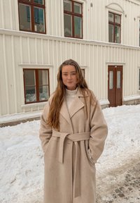 Νέα γυναίκα με μπεζ παλτό στέκεται έξω στο χιόνι μπροστά από ένα κρεμ κτίριο με πολλαπλά παράθυρα και πόρτες με καφέ πλαίσια.