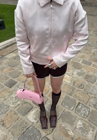 Chaqueta rosa claro con cremallera y mangas largas, pantalones cortos marrones, bolso rosa, medias negras transparentes y zapatos marrones de punta cuadrada sobre adoquines.