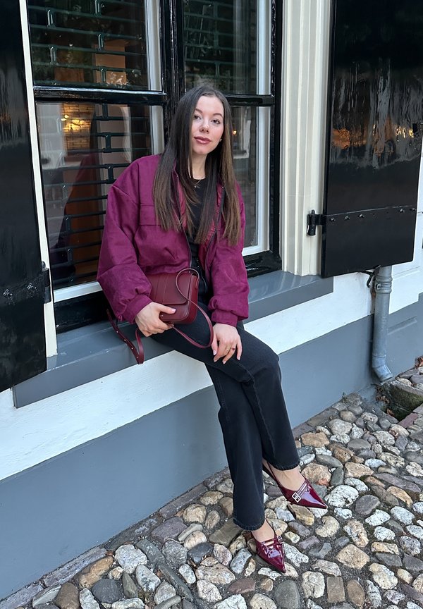 maroone Wildlederjacke, schwarzes Oberteil, dunkle Jeans und bordeauxfarbene Absatzschuhe mit einer Schnalle. Sitzt auf einem Steinweg neben einem Fenster mit schwarzen Fensterläden.