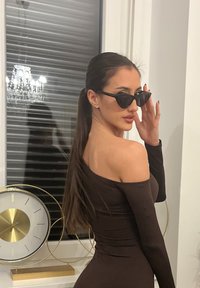 Femme avec une longue queue de cheval portant une robe noire à épaules dénudées et des lunettes de soleil œil de chat, posant à l'intérieur près d'une fenêtre avec une horloge décorative.