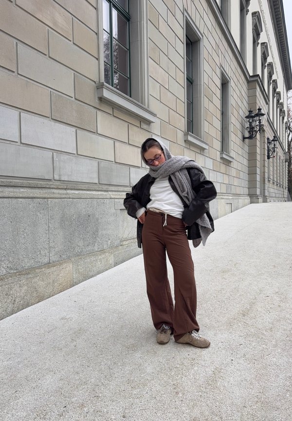 Person mit Brille, weißem Hemd, brauner Hose, schwarzer Jacke und grauem Schal steht auf hellem Steinpflaster in der Nähe eines hohen beigen Gebäudes.