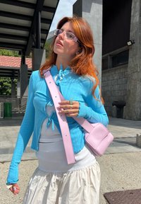 Ανοιχτό μπλε ριμπ κανελόνι με κορδόνια, φορεμένο πάνω από μια λευκή μπλούζα και μια ρέουσα λευκή φούστα; κρατώντας μια μικρή ροζ τσάντα χιαστί.