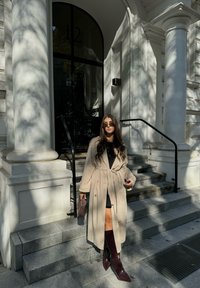 Μπεζ παλτό με ζώνη, μαύρη μπλούζα και μπορντό μπότες μέχρι το γόνατο; στέκεται σε πέτρινες σκάλες στο ηλιακό φως με σκιές από κοντινά δέντρα.