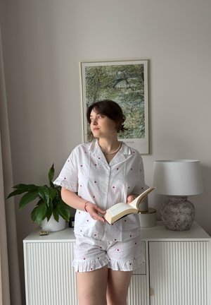 Persona con pijama blanco con pequeños corazones rojos está de pie leyendo un libro en una habitación con una planta, una lámpara y una obra de arte enmarcada de la naturaleza.