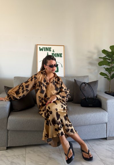 Mujer con un vestido largo de estampado y gafas de sol, sentada en un sofá gris con cojines negros, junto a un bolso negro y una planta verde en maceta.