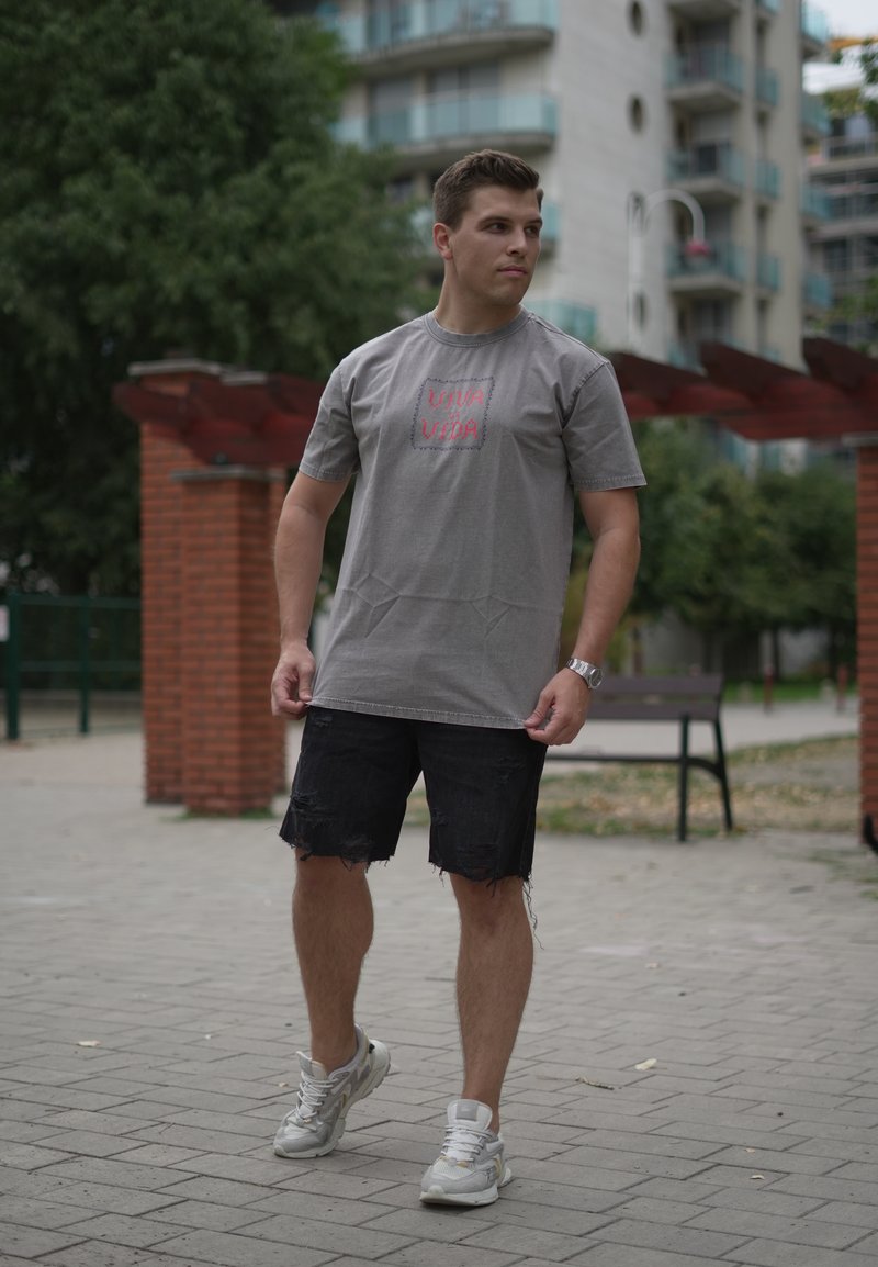Jeune homme debout à l'extérieur sur un chemin pavé, portant un t-shirt gris, un short noir déchiré, des baskets blanches et une montre-bracelet.
