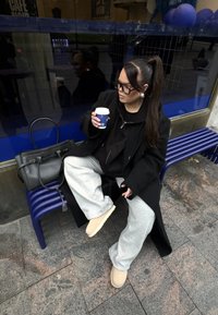 Sort frakke, grå joggingbukser og beige sko båret af en person, der sidder på en bl�å bænk og holder en kaffekop, med en taske ved siden af.