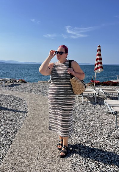 Vestido maxi sin mangas a rayas con un patrón en blanco y negro; accesorio con gafas de sol negras y un bolso de mimbre; de pie junto al agua.