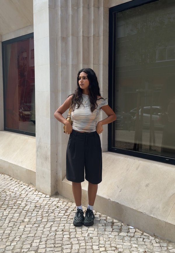 Joven mujer con el pelo largo y oscuro está de pie en una acera de adoquines cerca de un edificio de piedra, vestida con una camiseta blanca, pantalones cortos negros y zapatillas negras.