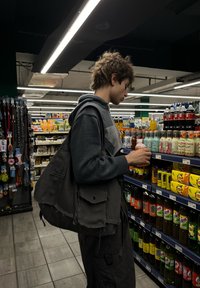 Žmogus su garbanotais plaukais, vilkintis pilką gobtuvą ir tamsias krovinines kelnes, laiko gėrimą maisto prekių parduotuvės eileje tarp spalvingų gėrimų produktų.