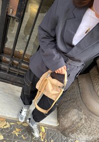 Personne portant un blazer gris et un pantalon noir tenant un sac à main beige debout sur un trottoir couvert de feuilles tombées près d'une grille métallique.