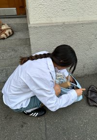 Helles blaues Tragetasche auf dem Boden mit sichtbaren Inhalten, darunter ein brauner Zylinder. Weißes Shirt und Jeansshorts getragen. Schwarze Schuhe mit weißen Streifen.