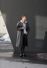 Szary wełniany płaszcz, szara bluza z kapturem, czarne spodnie i czarne botki; stojący na mokrym chodniku z geometrycznym ciemnym tłem.