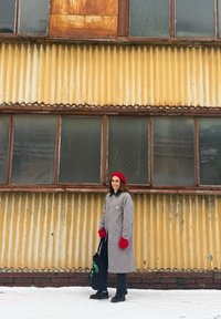 Femme portant un béret rouge et des gants, manteau gris, tenant un sac fourre-tout noir, debout sur la neige devant un mur ondulé jaune rouillé avec des fenêtres.