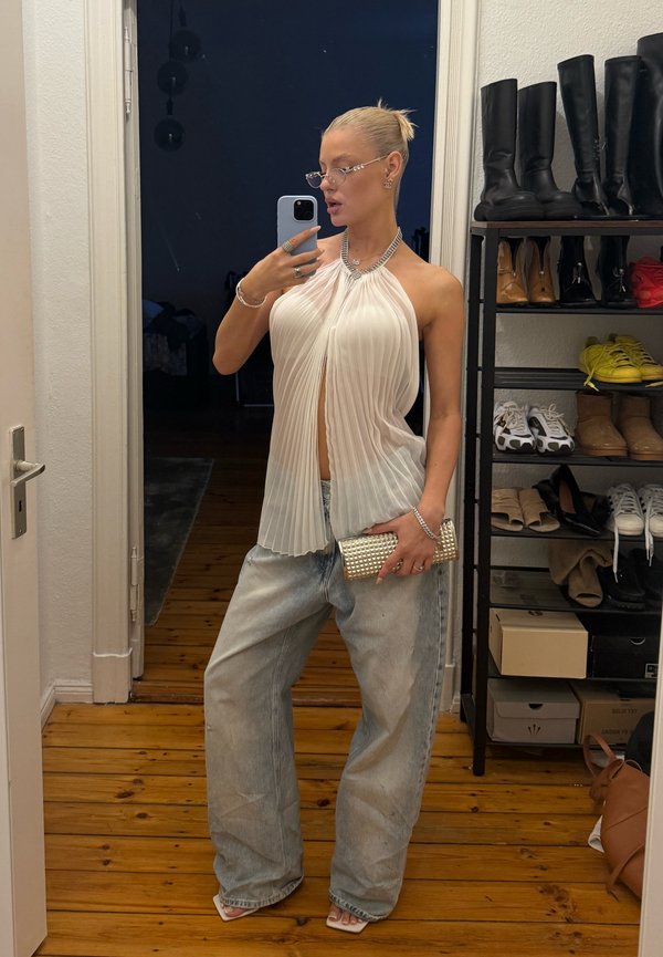 Vit plisserad halterneck-top, lösa blå denimjeans, klara bågar glasögon, silverkedja, och en metallisk clutch. Trägolv i bakgrunden.