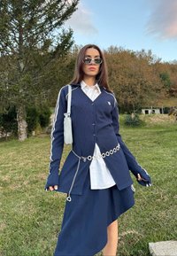 Femme portant une jupe asymétrique bleu marine et un cardigan avec une chemise blanche, lunettes de soleil, ceinture chaîne argentée, debout dehors sur l'herbe avec des arbres en arrière-plan.