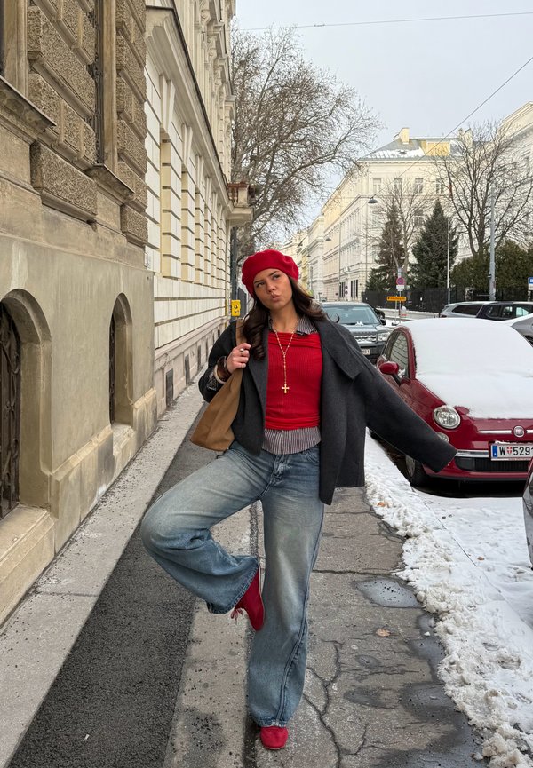 Mladá žena v červenom baretke a svetri pózuje na zasneženom mestskom chodníku, zdvihnutú jednu nohu, má na sebe sivý kabát, džínsy a červené topánky, nesie hnedú tašku.