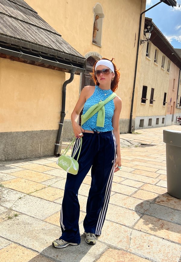 Crop top blu textured, pantaloni sportivi blu navy con strisce bianche, borsa verde, occhiali da sole e fascia bianca; in piedi su pavimentazione in pietra.