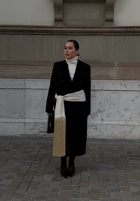 Lang sort dobbeltradet frakke med en hvid rullekrave, bundet med et hvidt frynsebælte. Sorte hæle og en sort håndtaske fuldender outfittet.